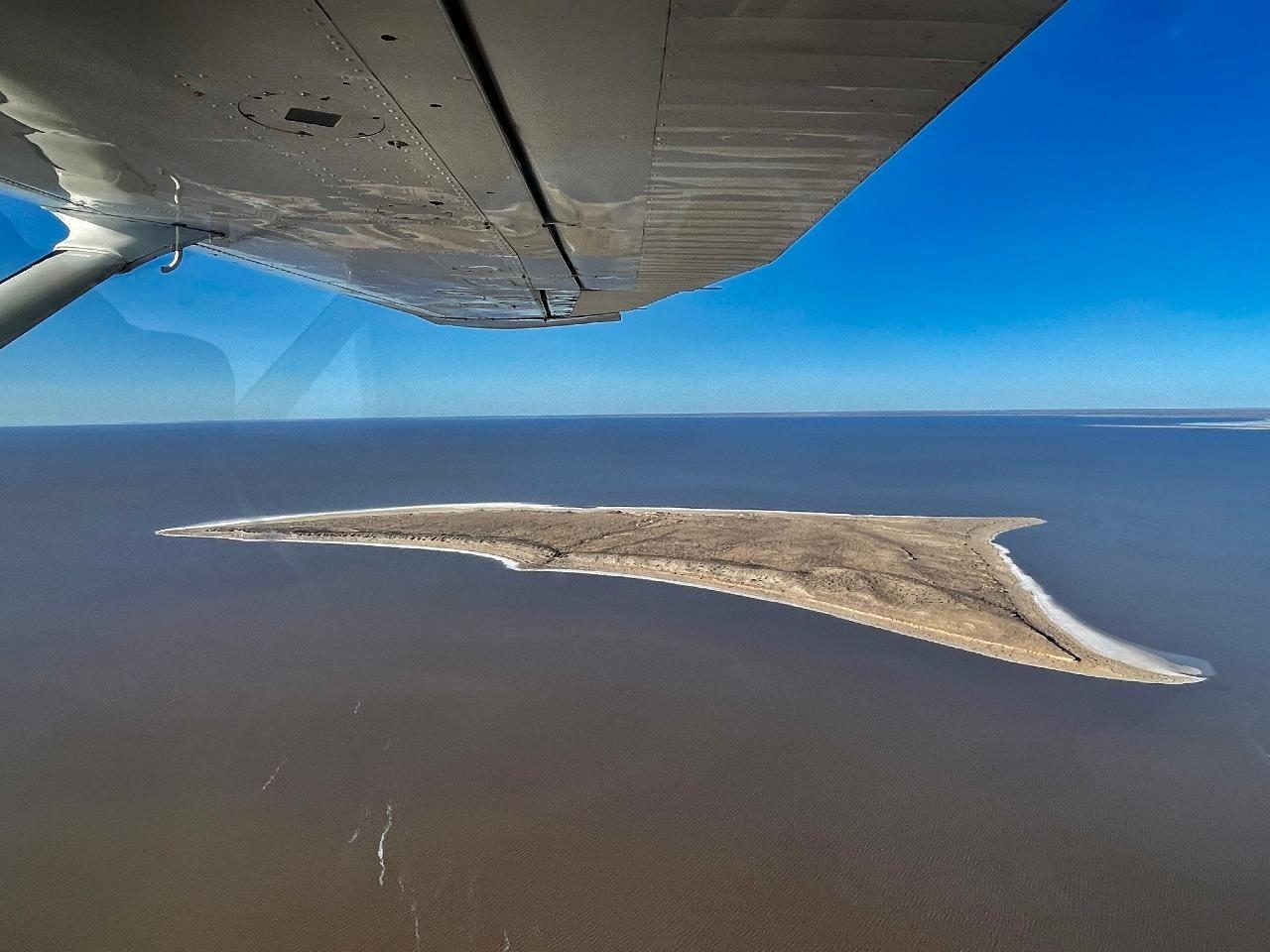 A plane wing above a sandy island in the blue water of Lake Eyre