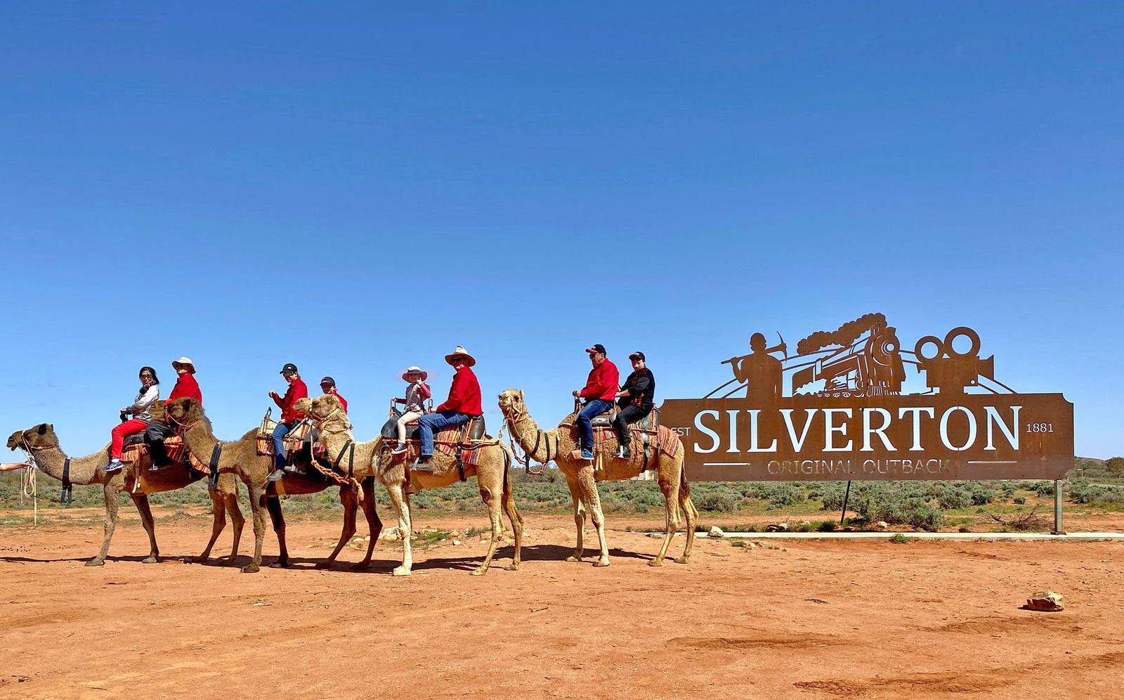 Camel riders passing the Silverton sign under a blue sky