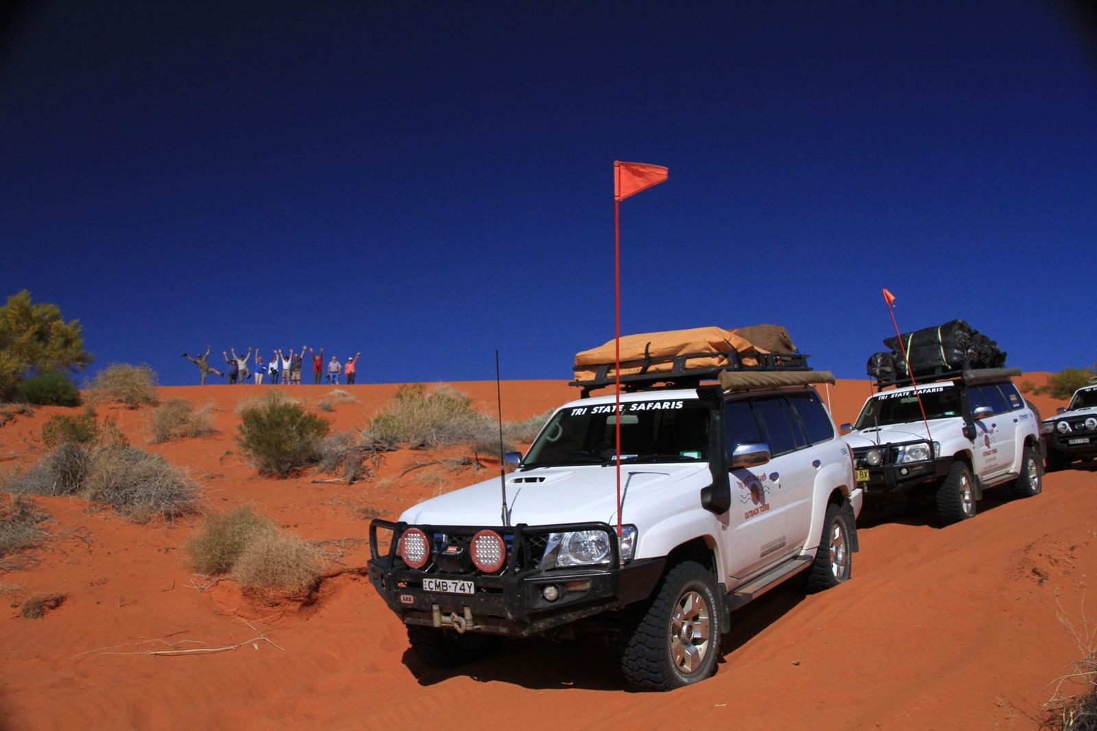 Two white Tri State Safaris 4WDs on a red dune under a deep blue sky, with guests waving from the crest